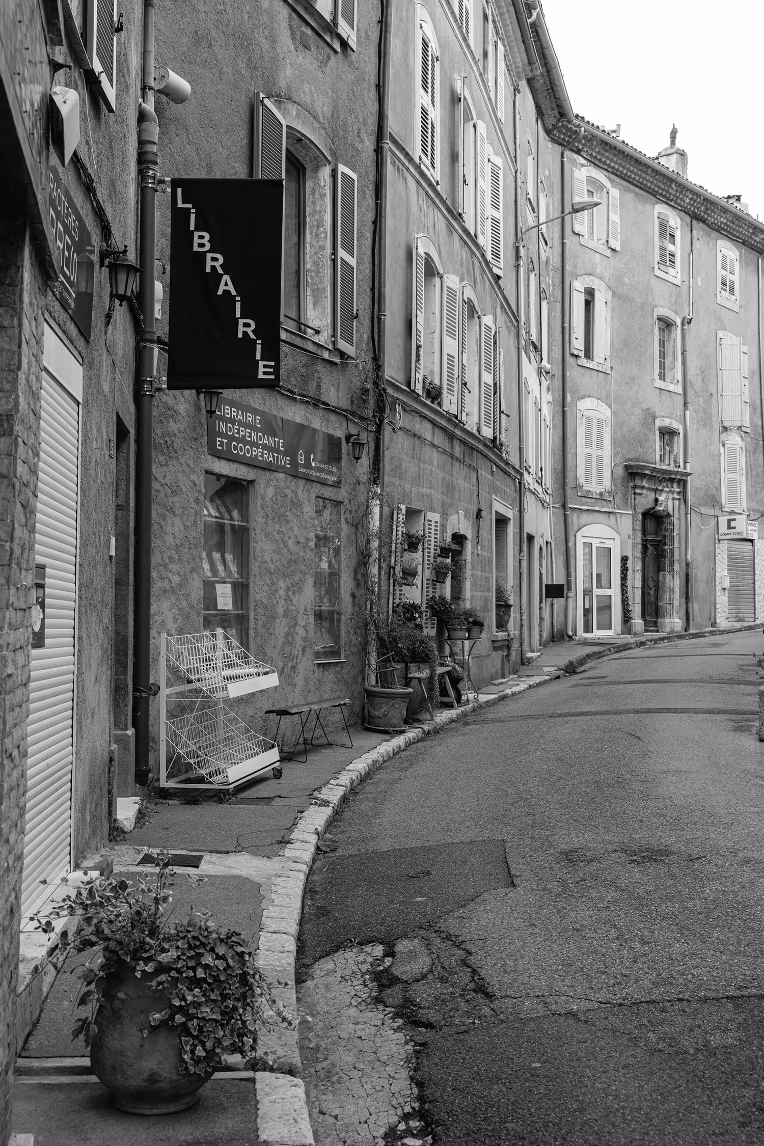 Atmospheric black and white street photography in Aups, Provence, France, showing a curved, narrow street lined with traditional French buildings with closed shutters, featuring a prominent 'Librairie' sign and an overturned market cart.