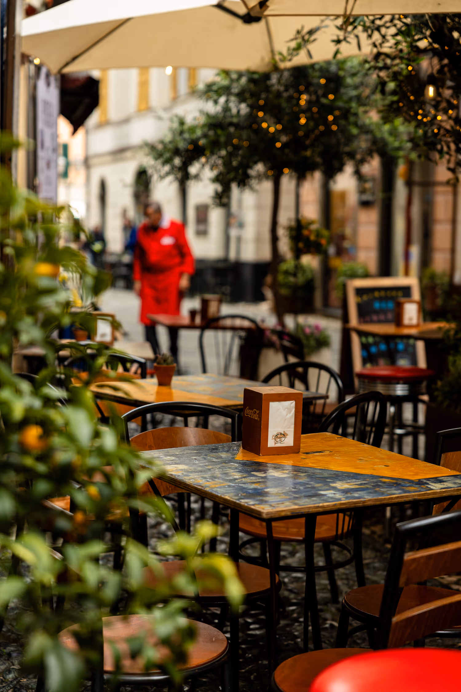 Atmospheric view of a narrow, cobbled alley cafe in Finalborgo, Italy, with empty wooden tables and a figure in a red uniform blurred in the background, emphasizing shallow depth of field (bokeh).