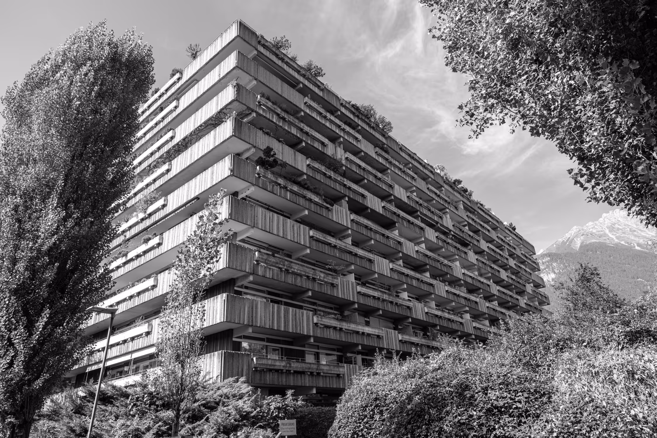 Low-angle, monochrome view of Franz Kotek's brutalist residential building in Innsbruck, Mariahilfpark, framed by trees, emphasizing repetitive geometric balconies and the building's massive scale against a mountainous backdrop.
