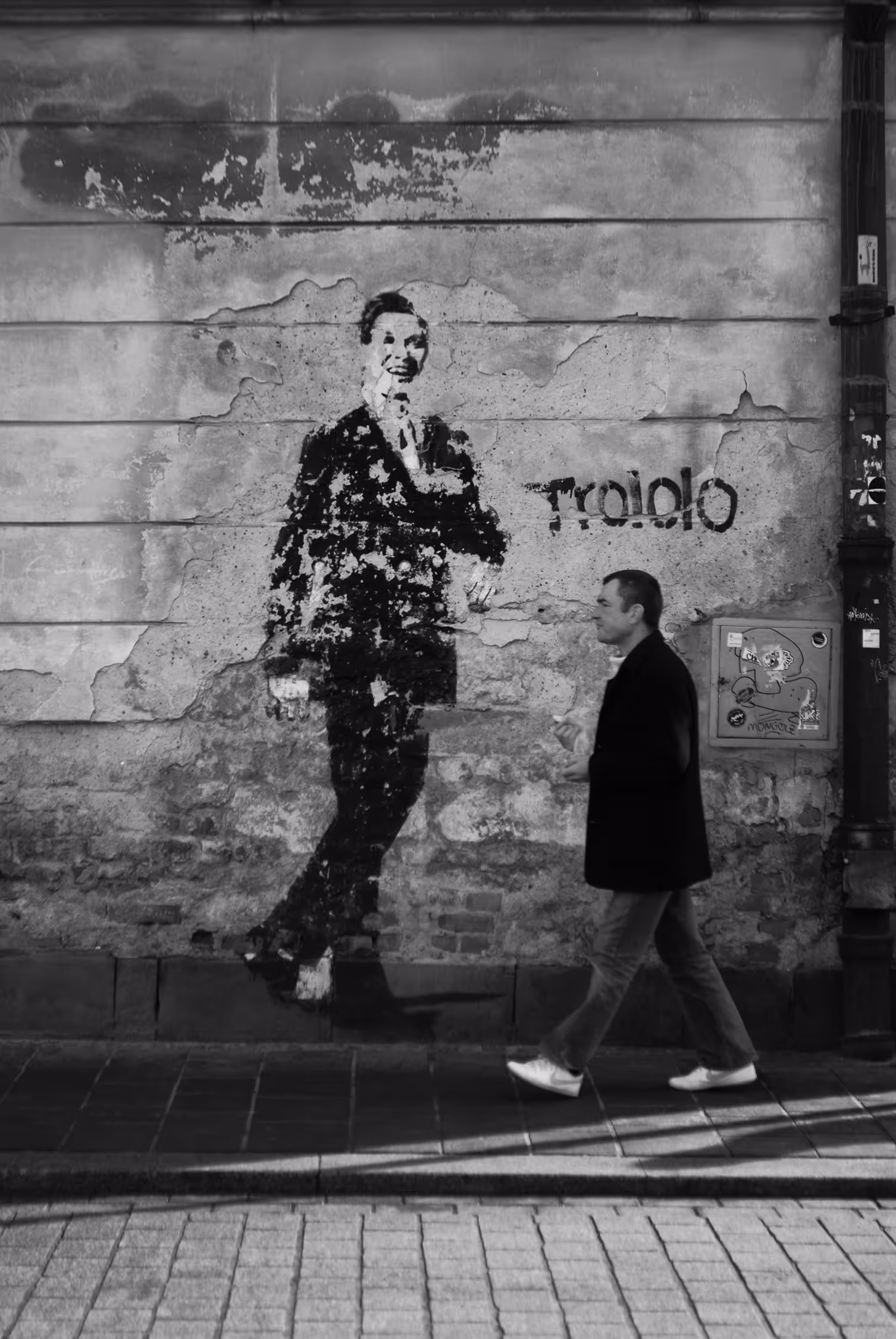High-contrast, black and white street photograph in Kraków, Poland, showing a pedestrian walking past a dilapidated wall with a large mural of the singer Eduard Khil ("Trololo"), emphasizing the contrast between the walking figure and the street art.