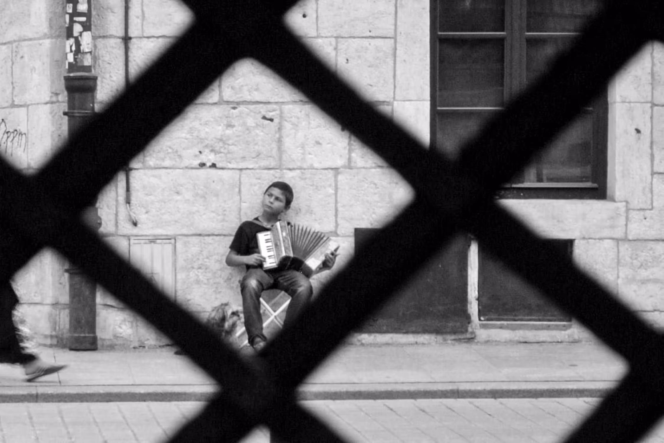Black and white candid street photography in Krakow, showing a young musician playing an accordion behind a dramatic, diamond-shaped grid foreground, framed by an old stone building wall.