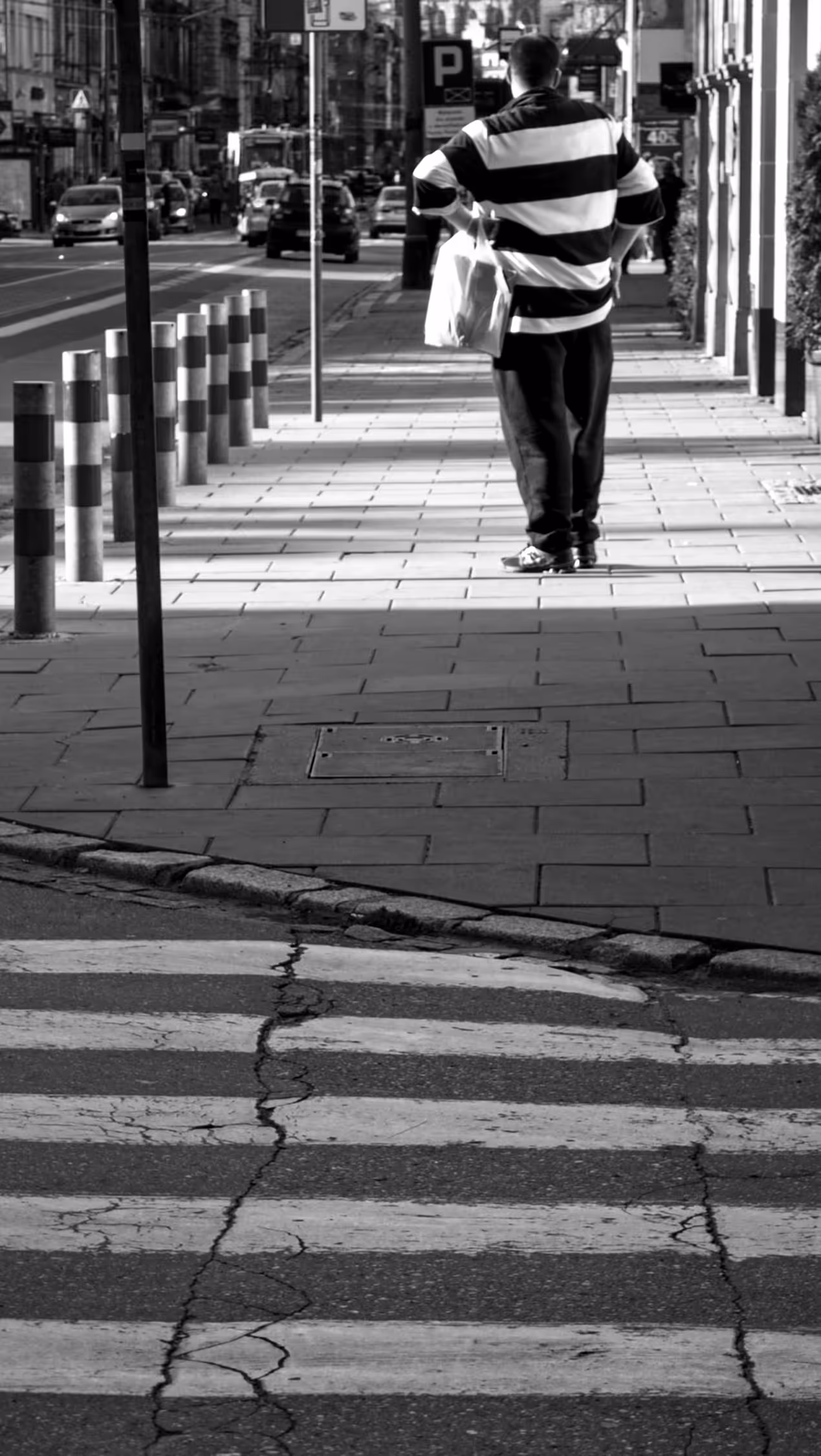 High-contrast black and white street photography in Krakow featuring a candid shot of a man in a striped shirt standing on a sunlit sidewalk, creating a geometric composition with urban textures and shadows.