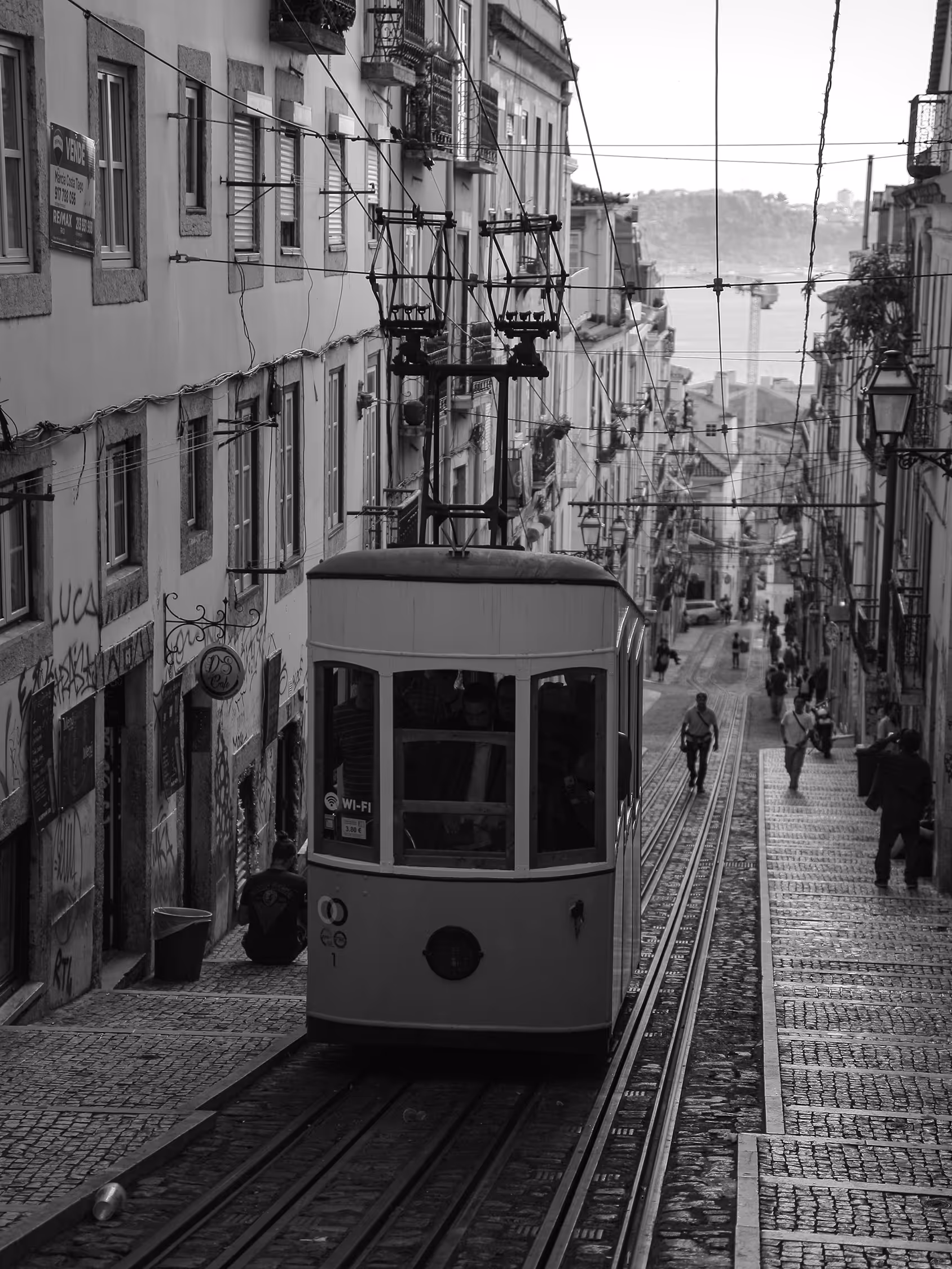 Atmospheric black and white street photography of the Elevador da Glória funicular ascending a steep, narrow cobblestone street in Lisbon, Portugal, framed by tall, traditional residential buildings, emphasizing depth and historical urban texture.