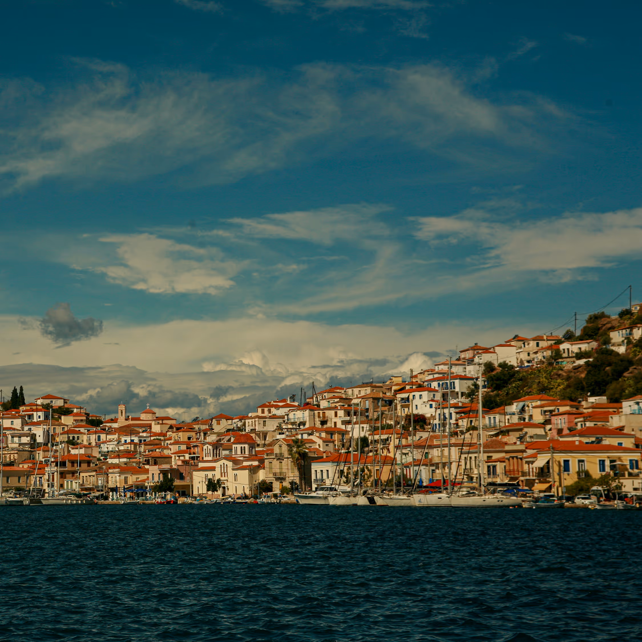 A vibrant, scenic view of Poros island harbor in Greece, featuring white houses with terracotta roofs cascading down the hill behind moored sailboats, set against a deep blue sea and dramatic sky.