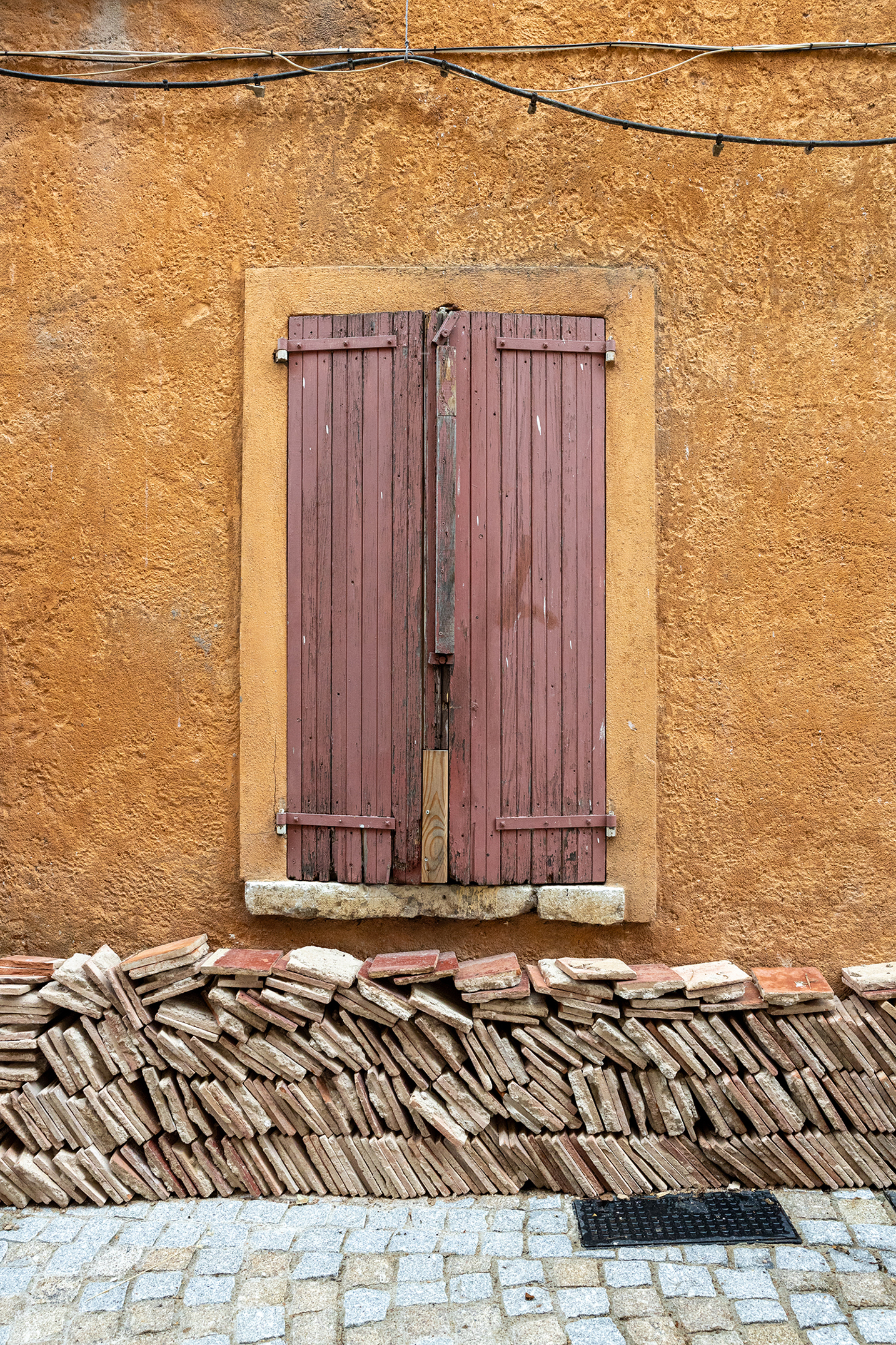 Vertical photograph in Sainte-Croix-de-Verdon of weathered red wooden shutters on a vibrant, textured ochre wall, with a stack of old terracotta roof tiles forming a base, emphasizing texture and symmetry.